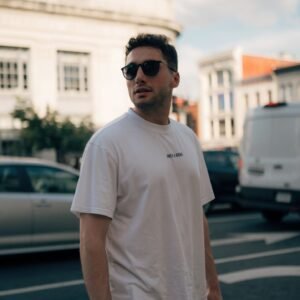 Stylish man wearing sunglasses standing on a city street with urban backdrop.