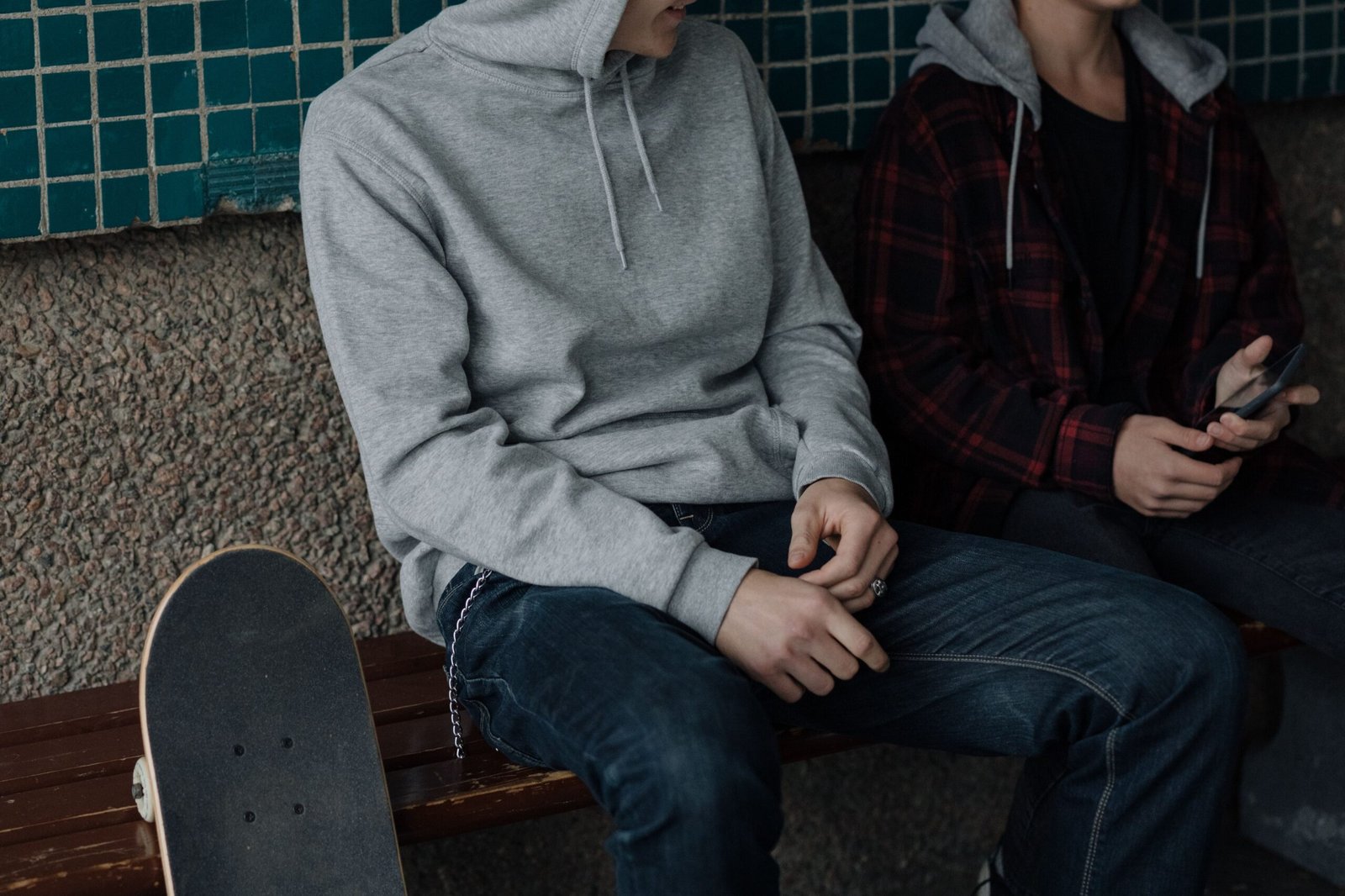 Two teenagers sitting on a bench with a skateboard, wearing casual hoodies in an urban environment.